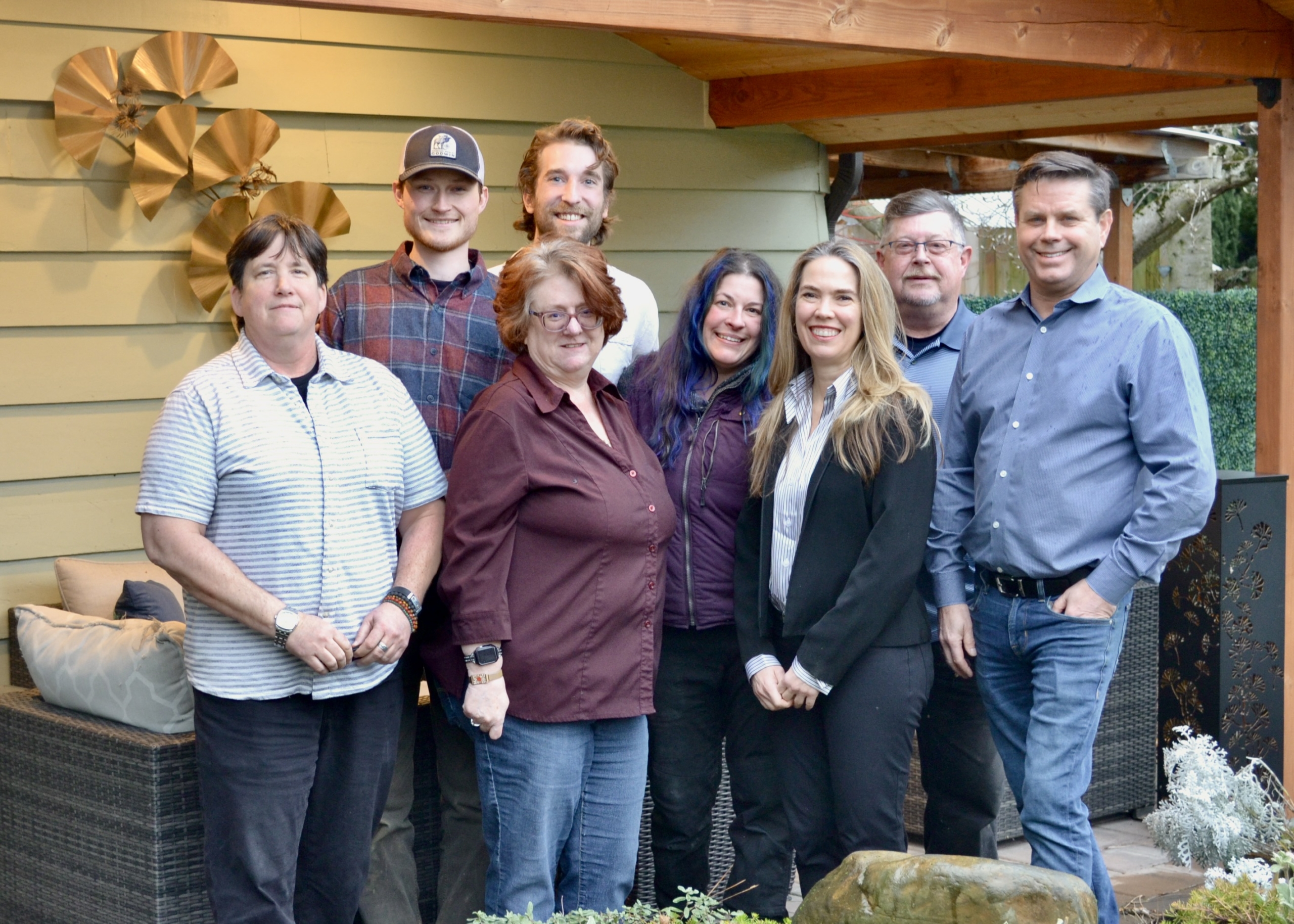 DSC_0347 Eight adults stand together in a backyard patio area, posing for a group photo in front of a beige wall and outdoor seating at the home of a Portland Sustainable Home Builder.