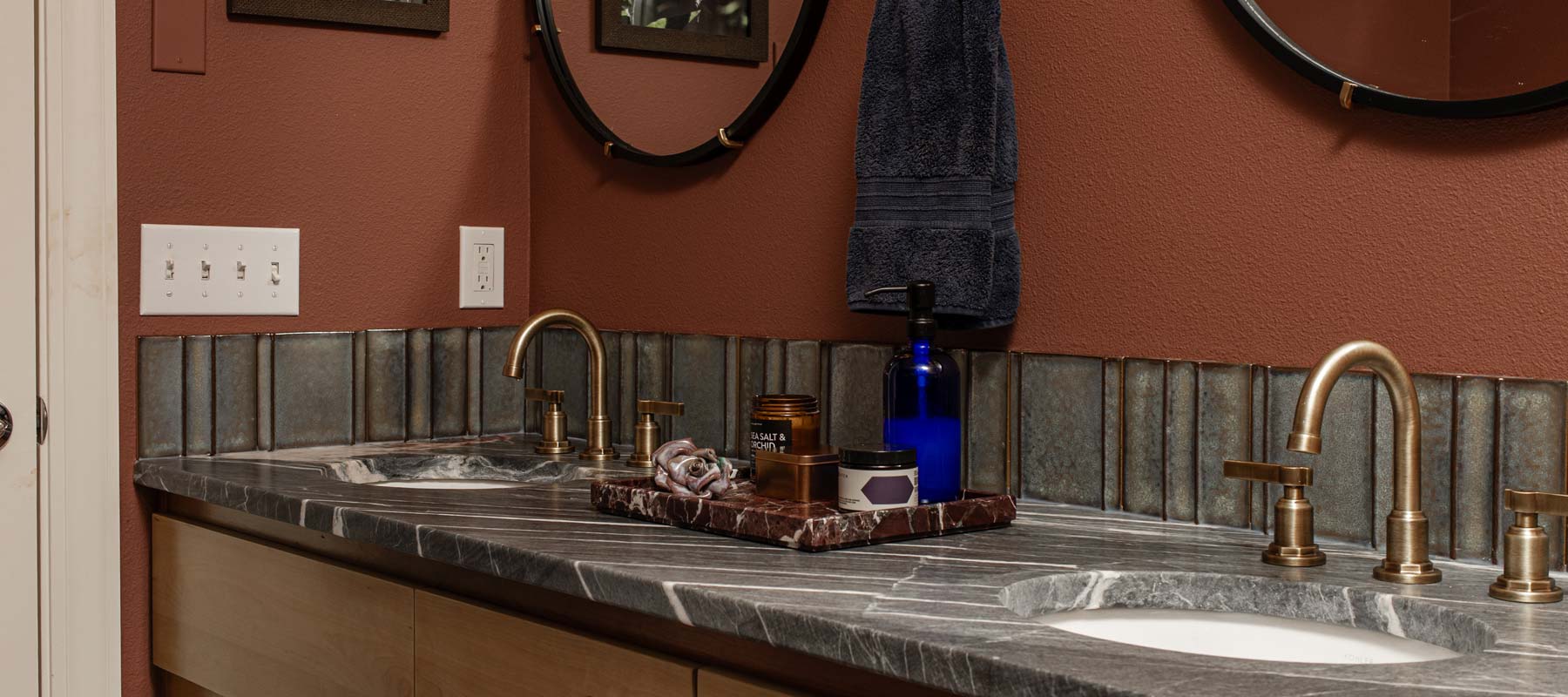 Double bathroom sink with brass faucets, gray marble countertop, decorative tray with toiletries, dark towel, and two round mirrors against a brown wall.