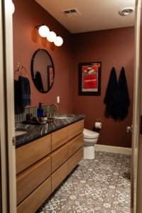 Modern bathroom with a dark countertop, wooden drawers, patterned tile floor, round mirror, wall art, and navy towels on hooks. The lighting features three globe bulbs above the mirror.
