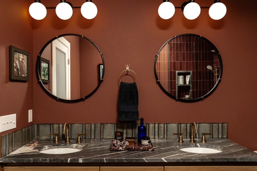 Double-sink bathroom with two round mirrors, marble countertop, brass faucets, dark towel, and wall-mounted lights above each mirror.