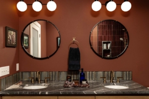 Double-sink bathroom with two round mirrors, marble countertop, brass faucets, dark towel, and wall-mounted lights above each mirror.