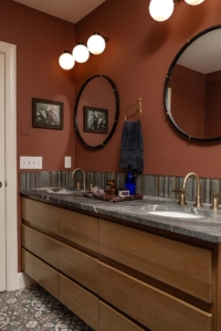 Double-sink bathroom with wooden cabinets, gray countertops, two round mirrors, gold fixtures, wall art, and globe lights against a rust-colored wall.