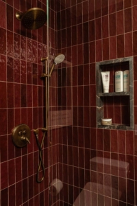 A shower with dark red tile walls, brass fixtures, a round overhead showerhead, a handheld showerhead, and a built-in shelf holding three bottles and a round container.