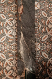 A vertical slab of dark marble with white veins stands against a detailed geometric mosaic tile wall in earthy tones.