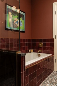 A bathroom with dark red tiled bathtub, brass fixtures, brown walls, patterned floor, and a framed “Hampton Library” art print above the tub.