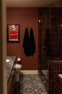 Bathroom with patterned tile floor, brown walls, two dark towels, a wall poster, a toilet, marble countertop, and a glass-enclosed shower with dark tile and brass fixtures.