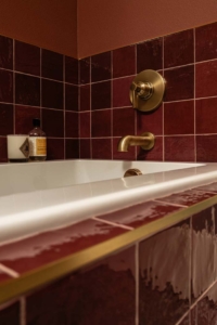 Close-up of a bathtub with red tiles, gold faucet, and wall-mounted controls. A bottle and cup are placed on the edge.