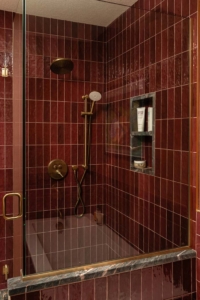 Shower with maroon vertical tiles, brass fixtures, built-in shelf holding toiletries, and a glass door with a brass handle.