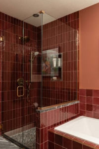 Modern bathroom with a glass-enclosed shower, red tiles, built-in shelf with toiletries, and a white bathtub with a dark countertop edge.